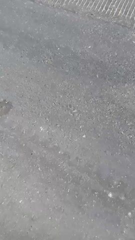 The video displays a person walking on a paved surface. The surface has visible texture and some debris. As the video progresses, the camera angle shifts, revealing a wider outdoor environment with a clear sky and some distant structures. A white car is visible in the distance, moving along a road.