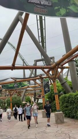 The video shows a bustling scene within an amusement park, specifically the Jurassic World themed area, likely at Universal Studios Japan in Osaka. Numerous people, including families and groups of friends, are casually walking along a paved pathway. Above them, towering grey rollercoaster tracks weave through a large, supportive framework of what appears to be a protective canopy or net, suggesting an indoor or covered outdoor section of the ride.

Along the path, large decorative structures resembling bamboo or rustic wood, lashed together, create a jungle-like archway. Banners displaying the "JURASSIC WORLD" logo with dinosaur silhouettes hang from these structures, reinforcing the theme. Lush green foliage and bushes line the path, adding to the prehistoric jungle atmosphere.

The weather appears to be overcast during the daytime, but people are dressed in light casual wear, indicating a comfortable temperature. Many individuals are sporting novelty headwear, such as animal ears or dinosaur-themed hats, commonly seen in theme parks. People are generally relaxed, walking at a leisurely pace, with some looking around at their surroundings. The overall impression is one of an enjoyable day at a popular attraction.