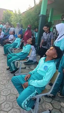 The video shows a group of children, mostly boys wearing teal uniforms, sitting on chairs outdoors. They appear to be participating in a game where they have biscuits balanced on their foreheads. Some children are looking up with their mouths open, seemingly trying to eat the biscuits without using their hands. One boy in the foreground has a biscuit on his forehead and is looking up with his mouth open. In the background, other children and adults are watching, some smiling and some appearing to be cheering. The environment looks like a schoolyard, with trees and a building visible. The time stamp indicates the video is very short, just four seconds long.