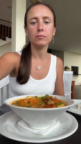 A woman with long brown hair, tied back in a ponytail, is seated at a table, enjoying a meal. She is wearing a white tank top and a white, shell-like necklace, along with dangling earrings. In front of her is a large white bowl filled with a colorful dish that appears to be a stew or curry, containing green beans, orange chunks (possibly tomatoes or carrots), and other ingredients in a reddish-orange broth. The bowl rests on a white plate with a white napkin underneath.

The woman uses a silver spoon to scoop a portion of the food from the bowl. She brings the spoon to her mouth, tastes the food, and her expression shifts from neutral to a pleasant smile, indicating satisfaction with her meal. The background suggests an indoor dining setting, possibly a restaurant or a hotel dining area, with white chairs and tables visible, as well as a staircase to the left. There is background chatter, but the woman herself does not speak in the video.