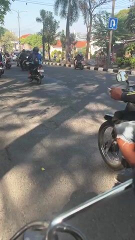 The video shows a street scene with multiple motorcycles. In the foreground, the perspective is from a motorcycle, with handlebars visible at the bottom. A motorcycle in the mid-ground has a rider wearing a yellow and black jacket and a black helmet, with two blue barrels strapped to the back. Another motorcycle with a rider in a red jacket is also visible. The background shows a road with other vehicles and trees. The lighting suggests it is daytime.