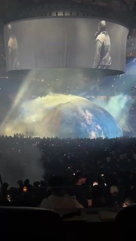 A stadium or arena is filled with a large crowd of people, mostly visible from behind. The lighting is dim, but many attendees are holding up their phones, presumably to record the performance. The stage is dominated by a large, rounded structure resembling Earth, partially covered in fog. On a massive circular screen suspended above the stage, black-and-white video of a person resembling Kanye West is projected, with the images mirrored on either side of the screen. There are bright lights illuminating the structure, adding to the visual spectacle of the concert.