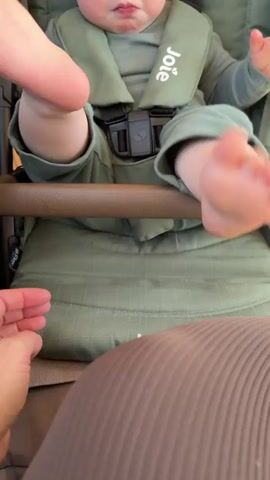 A close-up shot shows a baby in a green outfit strapped into a green stroller. The baby's bare feet and lower legs are visible, resting on the stroller's brown support bar. The baby's hands are also seen, reaching towards the bar. An adult hand gently touches one of the baby's feet. The adult, wearing a taupe-colored ribbed top, is partially visible in the lower part of the frame. The stroller has a "Joie" logo on the harness. The background is slightly blurred.
