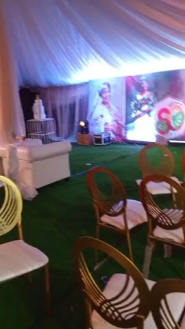 The video shows an event setup, likely a party or reception, taking place indoors under a white tent with blue lighting. The floor is covered in artificial green turf. In the foreground, there are several gold-colored chairs with white cushions. To the left, there is a white sofa and a tiered white cake on a white stand. In the background, there is a large screen displaying a portrait of an older woman smiling. Several stage lights and black equipment cases are also visible on the ground. The video is shaky and primarily focuses on the arrangement of the venue.