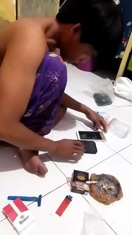 A person is sitting on the floor with their back to the camera, wearing purple patterned pants. They are leaning forward and looking down at two cell phones. In front of them on the floor are a pack of cigarettes, a lit cigarette in an ashtray, a lighter, and a razor. To their left is another pack of cigarettes and a blue object. A plastic bottle of water is to their right, and in the background, a pink container and other objects are visible. The floor appears to be tiled.