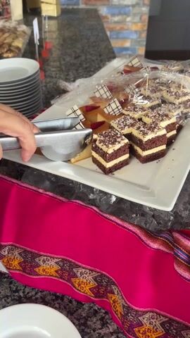The camera captures a person using metal tongs to select a piece of "Crema Volteada" (a type of custard dessert) from a buffet display. The person places the dessert on a white plate. The buffet features two types of desserts: crema volteada and small chocolate layered cakes. The desserts are arranged on a rectangular white plate atop a bed of ice, partially covered with clear plastic wrap. A colorful pink table runner with geometric patterns adorns the front of the buffet. In the background, there are stacks of white plates and a glimpse of other buffet items. The environment appears to be a hotel or restaurant breakfast buffet.