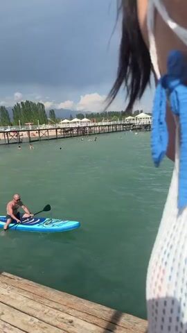 This media file depicts a sunny day at a beach in Sary-Oy, Kyrgyzstan. The scene is set on a wooden pier extending over clear, turquoise water. 

The primary subjects are people enjoying a day at the beach. The video begins with a close-up of a woman's back, wearing a white mesh cover-up over a blue bikini. The camera then pans out to reveal a wide shot of the sea and sky. We see several individuals swimming in the water, including a man in the distance.

A young woman in a bikini is shown at the edge of the pier, preparing to jump into the water. She appears excited and is looking towards the sea. Another young person, a boy wearing floaties on his arms and a baseball cap, is walking along the pier, also heading towards the water. Other people are visible further down the pier, some sitting and others standing.

The weather is clear and sunny with a bright blue sky dotted with a few white clouds. The sea is calm and inviting. The wooden pier is a prominent feature, showing signs of wear and tear, adding to the casual, beachside atmosphere.

The overall mood is one of leisure and fun, with people enjoying a summer day by the water. There is no visible text in the media file.