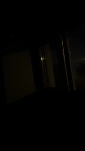 The video shows a dark room with a window. On the left side of the screen, there is a light-colored rectangular object, possibly a wall or a piece of furniture. In the center, there is a window divided into two vertical panels by a dark frame. Through the window, a dark outdoor scene can be seen, with faint lights in the distance and silhouetted trees against a dark sky. The lighting in the room is dim, with a faint light source visible inside the left panel of the window. The video is static.