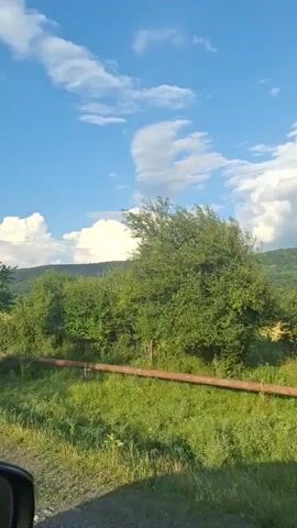 The video captures a dynamic landscape scene, likely filmed from a moving vehicle. The primary subjects are the lush green trees and vegetation that dominate the foreground and midground, with a clear blue sky dotted with fluffy white clouds overhead. The camera pans, offering glimpses of rolling hills and mountains in the distance, suggesting a rural or semi-rural setting. The time of day appears to be late afternoon or early evening, judging by the angle of the sunlight and the warm hues on the horizon. The weather seems pleasant, with a clear sky indicating fair conditions.

Several notable objects are visible, including what appears to be a long, reddish-brown pipe or log running horizontally across the middle ground of the frame, partially obscured by the grass and foliage. The trees vary in size and density, with some appearing as mature, spreading specimens and others as smaller shrubs.

The overall mood conveyed is one of natural beauty and tranquility. The movement of the camera suggests a journey or scenic drive through the countryside. While no people are visible, the presence of the pipe might hint at human activity or infrastructure in the area, though it is integrated into the natural landscape. The location context provided, Alkhasty, Russia, places this scene in a specific geographical region, likely characterized by such natural landscapes. There is no visible text in the media file.
