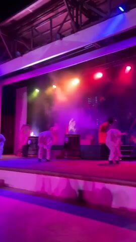 This is a medium shot video of an outdoor stage performance at night. The stage is illuminated by vibrant pink and purple lights on the audience side, and a mix of pink, green, and yellow lights on the stage. Smoke or fog drifts across the stage, catching the colored lights. Five individuals dressed in white, mummy-like costumes are performing a synchronized dance. Two figures in the background are dressed in red and black, one appearing to wear a black hat and the other a red dress. The stage has several wooden barrels and what appears to be a staircase on the left side. The performance seems to be of a spooky or theatrical nature.