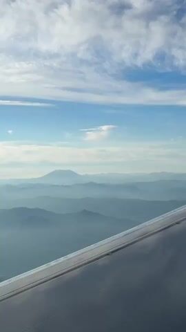 This video is a point-of-view shot from an airplane window, looking out over a landscape of mountains and clouds. The airplane's wing is visible in the foreground, extending from the bottom right of the frame. The wing is metallic and appears to be a dull gray color. The sky is blue with scattered white clouds, some of which are wispy and streaky. In the distance, several layers of mountains are visible, fading from a darker blue in the foreground to a lighter blue and gray in the distance. The furthest mountain is snow-capped and appears to be a volcano. The video pans slightly to the right, revealing more of the mountain range and the sky. The overall impression is one of vastness and natural beauty.