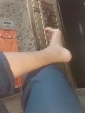 This is a close-up, low-angle video of a person's bare foot and lower leg. The person is wearing blue jeans. The foot is lifted slightly and the toes are visible. The background includes a wooden surface, brickwork, and an orange fabric. The lighting is dim.