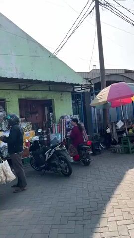 The video shows an outdoor market scene. There are several motorcycles parked on a paved area in front of a building with a light green facade. The building appears to be a small shop or stall selling various goods, including what looks like textiles or bedding. Several people are interacting in front of the stall. One person, wearing a black hijab and black clothing, is standing near a motorcycle, looking at items displayed. Another person, wearing a red shirt and a helmet, is holding out some items to the person in the hijab. To the right, two more individuals are seen near a motorcycle, one wearing a dark jacket and the other a maroon top, appearing to be engaged in a transaction or conversation. The sky is visible in the background, and power lines run overhead. The overall atmosphere suggests a busy marketplace.