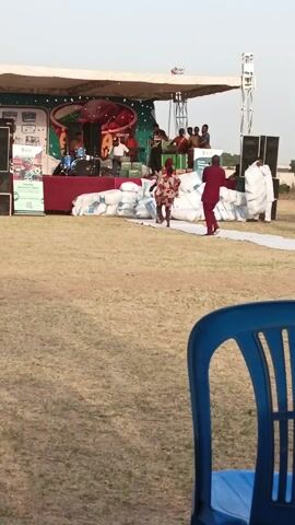The video shows an outdoor event with a stage set up on a grassy field. On the stage, a band is playing, and there are several people standing around. In front of the stage are stacks of large white bags and a white runner. Two people in colorful outfits are dancing on the runner, while a man in a red suit walks nearby. In the foreground, there is a blue plastic chair and the arm of a person wearing a red dress. The camera appears to be held by someone sitting in a chair, as the arm is visible. Other people can be seen standing around the stage. The sky is bright, indicating it is likely daytime.