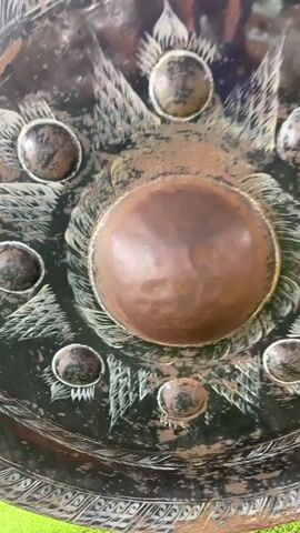 The video shows a close-up view of an ornate, circular object, possibly a shield or decorative plate, with a dark green background and raised copper-colored circular elements. The object is decorated with etched designs that appear to be stylized leaves or feathers. A hand, adorned with a turquoise beaded bracelet, enters the frame and gently touches the largest central copper element. The hand moves across the surface, highlighting its texture. The camera remains focused on this central area, with a slight blur in the background.