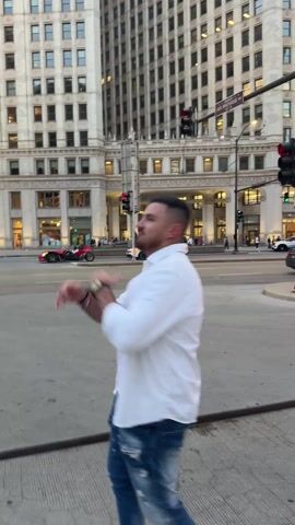 A man standing on a city sidewalk is shown from a low-angle shot. He is wearing a white button-down shirt, a white t-shirt, and distressed jeans. He appears to be clapping his hands, smiling, and moving in place. In the background, a red three-wheeled vehicle is seen driving across the street, in front of a Ghirardelli chocolate store. Buildings are visible in the background. A few people can be seen behind the parked car, and other cars are driving down the street.