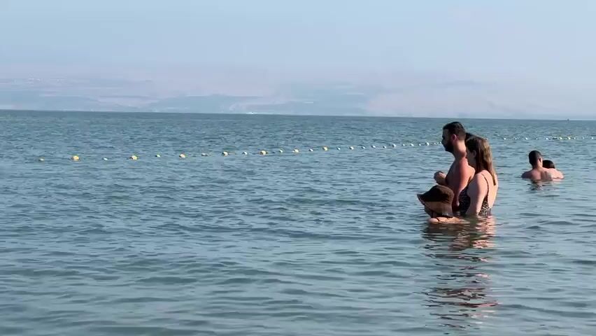 This video shows a body of water, likely a lake or sea, under a clear sky. In the foreground, three people are partially submerged in the water. Closer to the camera, a person wearing a wide-brimmed hat is visible, with their head and shoulders above the water. To their right, a couple stands in the water. The man is wearing dark shorts and the woman is wearing a patterned swimsuit. The man has his arm around the woman. Further in the background, another person is swimming. A line of yellow buoys stretches across the water in the mid-ground. In the far distance, a hazy landscape of hills or mountains is visible. The water is calm with gentle ripples. The overall atmosphere is serene.