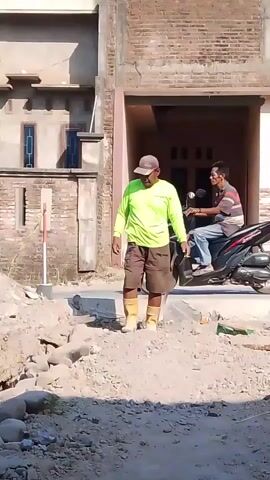 A man wearing a bright green long-sleeved shirt, brown shorts, and yellow boots stands in a dirt construction area. He is holding a shovel. A motorcycle with two people on it rides past him in the background. The man looks down towards the ground as he walks.