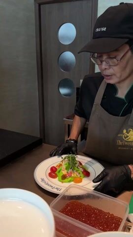 A chef, wearing a black cap with "CRUISE" written on it, glasses, a black shirt with green trim, and a brown apron with "The Twenty Eight HEDONIST SOCIETY" logo, is shown in a kitchen or food preparation area. They are wearing black gloves.

The chef is holding a white plate with a blue rim, which also bears the text "The Twenty Eight HEDONIST SOCIETY". On the plate is a meticulously prepared dish. The dish features a base of light green, possibly a puree or sauce, topped with shredded white meat, likely crab. Surrounding this are small red cherry tomatoes and a few yellow/orange cherry tomatoes. The dish is garnished with green leafy elements, resembling arugula or microgreens, and small orange spheres, which appear to be salmon roe or caviar. A lighter yellow/orange sauce or dressing is also visible on the plate.

In the foreground, to the right, there is a clear plastic container filled with a large quantity of red, spherical items, consistent with salmon roe (ikura). A white bowl is partially visible on the left side of the frame. The chef is looking intently at the dish, seemingly inspecting or presenting it. The background features a dark countertop and wooden doors with three circular frosted glass inserts.