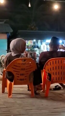 The video shows two people sitting in bright orange plastic chairs at night. They are facing away from the camera, towards a building with several lights on. The person on the left is wearing a light-colored headscarf and a greyish top. The person on the right is wearing a dark maroon top and has short hair. Both are seated at a table, though the table itself is not clearly visible.

The setting appears to be an outdoor area, possibly a roadside eatery or market stall in Keudeteupinpunti, Indonesia, given the provided location context. The ground is paved with brick-like stones. In the background, there are several motorcycles parked, suggesting a casual, possibly busy, environment. Above, string lights are hung, casting a warm glow and illuminating the scene, indicating it's nighttime. The overall atmosphere is relaxed and intimate, with the two individuals appearing to be engaged in conversation or sharing a meal. There are no discernible emotions conveyed from their posture alone, but the shared seating arrangement suggests companionship. No visible text is present in the frame.