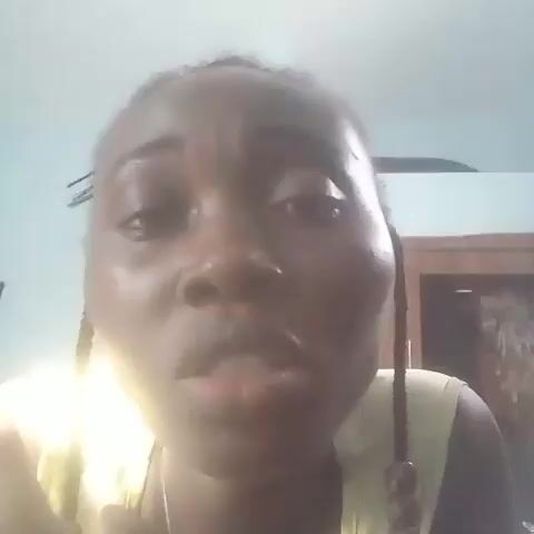 A close-up, low-angle, slightly out-of-focus selfie video features a young Black female with braided hair. She wears a yellow tank top and a necklace. Her eyes are wide, and she is looking directly at the camera. Her mouth is open and then closes slightly. Her right hand is raised, and her fingers are touching her chin. The background is a light blue wall with some indistinct objects on a shelf.