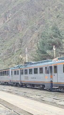 The video shows a silver train with an orange stripe running along the bottom of the passenger cars. The words "Machu Picchu Train" are visible on one of the train cars, and "First Class" is written on another. The train is labeled with the number 903. The train is on tracks in front of a hillside with a mix of green trees and tan, rocky terrain. There is a utility pole visible in the background.