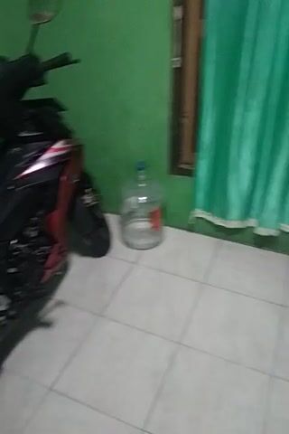 The video shows a motorcycle parked indoors. The motorcycle is black and red with a sporty design. It appears to be in good condition. Behind the motorcycle, there is a green curtain and a large plastic water bottle. The floor is tiled. There are no people visible in the video. The camera is held by hand and the footage is somewhat shaky, suggesting it was filmed by someone walking around the motorcycle.