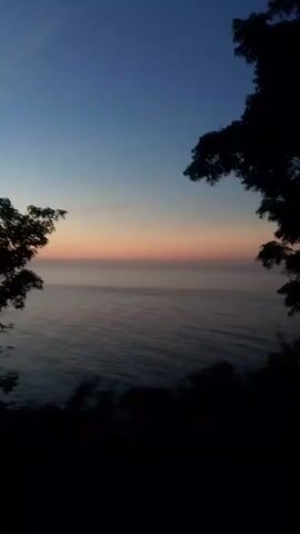 A video shows a wide shot of an ocean during twilight. The sky transitions from a deep blue at the top to a pale orange and pink band near the horizon. The ocean is dark and appears calm with subtle waves. Silhouetted trees frame the left and right sides of the shot, with the foreground also being dark and indistinct, suggesting a high vantage point overlooking the water. The scene is peaceful and quiet, with no visible people or activity.