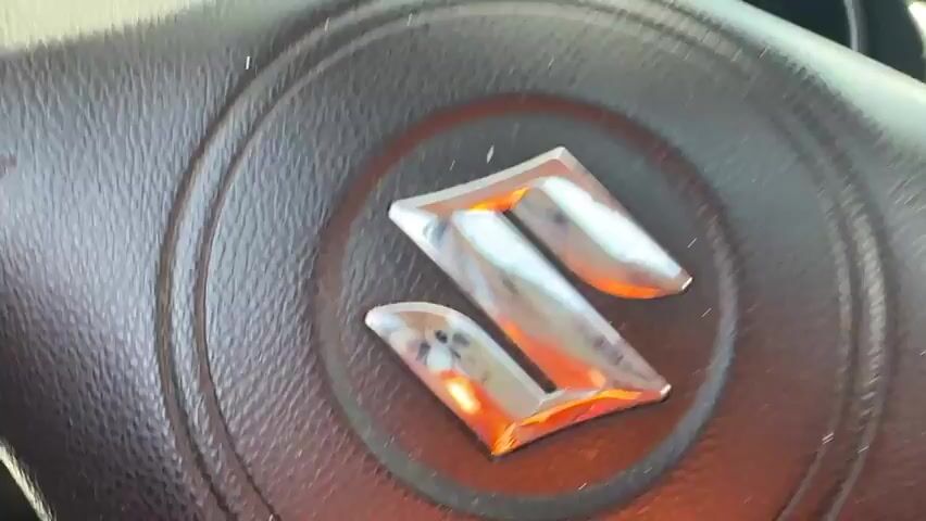 The video begins with a close-up of a car's dashboard and steering wheel, showing the Suzuki logo. The camera then pans out to reveal the road ahead. A speedometer shows the car is traveling at approximately 80 km/h. The road is a two-lane highway with a median barrier. On the right side of the road, there are billboards, one advertising "efbet" and another with a speed limit sign. Mountains are visible in the distance. The sky is blue with scattered clouds. The car passes other vehicles, including a white truck. The video continues to show the car driving on the highway, with the scenery of mountains and billboards passing by.