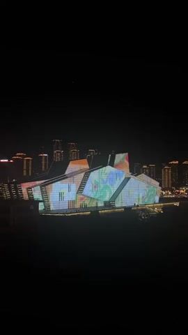 This video is a night-time shot of a large, modern building with a complex, angular structure. The facade of the building is covered in a dynamic, colorful light display that appears to be projected onto the surfaces. The lights shift and change, creating abstract patterns and shapes in various hues of green, blue, pink, orange, and yellow. The building is situated in an urban environment, as evidenced by the illuminated city skyline visible in the background, with numerous buildings and lights stretching into the distance. The foreground and immediate surroundings of the building are dark, suggesting the vantage point is from a distance or an elevated position. The overall impression is of an architectural structure transformed into a canvas for a vibrant light show.