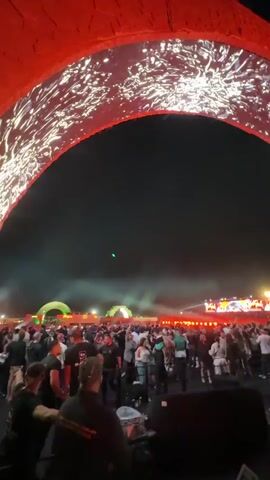 The video shows a crowded outdoor festival at night, with a large arched structure lit up with bright, swirling patterns. The camera moves erratically through the crowd, briefly capturing people dancing and enjoying the music. Later, the camera focuses on a woman in a white shirt who is standing on a staircase and appears to be recording a video of herself.