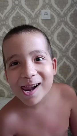 A young boy with a very short haircut is looking directly at the camera. He is shirtless, with his shoulders and chest visible. His skin appears to be tanned. The boy's facial expression changes slightly throughout the video. Initially, he has a puckered expression, then he smiles broadly, revealing his teeth. He then smiles with closed lips, and finally, his expression relaxes into a neutral, but still pleasant, look.

The background is a wall with patterned wallpaper. The wallpaper has a repeated damask or paisley design in muted colors of grey and beige. There is a white electrical outlet visible on the wall behind and slightly to the right of the boy's head. The lighting suggests it is daytime, and the setting appears to be indoors, possibly a bedroom, given the glimpse of a patterned bedspread or sheet to the left of the frame. There is no visible text in the media file. The boy's expression suggests a playful or engaging mood.