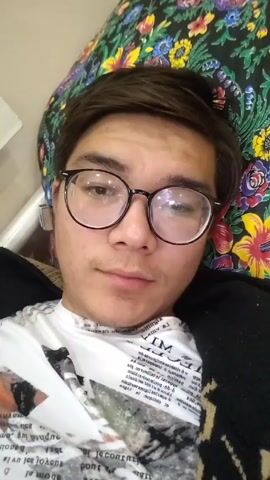 A young man wearing glasses, a white t-shirt with writing on it, and a black cardigan lies against a colorful floral pillow. He raises his hand in a wave. The scene is brightly lit.