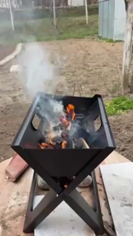 The video shows a small, black, diamond-shaped brazier burning wood and charcoal, with smoke rising in a yard. The brazier is elevated on a cross-shaped metal stand, sitting on a rough wooden surface alongside a brick and a stone slab. In the background, there's a fenced-off garden plot, some greenery, and a shed.
