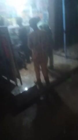 The video shows a dimly lit store, seemingly at night. Several people are inside, their forms indistinct but appearing to be engaged in casual conversation. One person is standing still, facing the store's interior, while others seem to be moving around. The store's interior is illuminated, displaying what appears to be shelves with products. The lighting is poor, creating a grainy and unclear image.