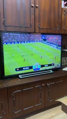 The video shows a television screen displaying a soccer match between Belgium and Egypt. The score is 0-0, and the time is 00:14. The game is being played on a green field with players in white and red uniforms. The television is placed on a wooden cabinet with ornate carvings. The background appears to be a room with wooden cabinets on the wall.