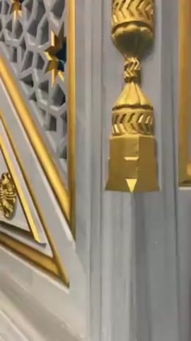 The video shows a low-angle, panning view of the interior of what appears to be a mosque or other religious building. The walls and staircase are decorated in a pale grey color with gold accents, including geometric patterns, stars, and Arabic calligraphy. The camera moves from close-ups of decorative elements to wider shots showing the overall design and architecture of the space, including the curved ceiling with embedded lights. There are no people visible in the video. The overall impression is one of ornate and detailed craftsmanship.