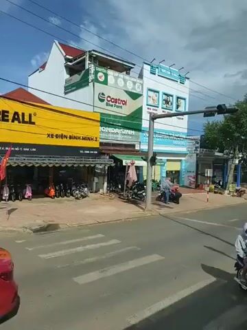 A street scene in Vietnam unfolds under a partly cloudy sky. The camera pans from right to left, showcasing a bustling commercial area. In the foreground, two individuals on a scooter, wearing masks and protective eyewear, are captured mid-motion. To the right, a white SUV is visible, and further ahead, a red and white circular "No Entry" sign stands prominently. Buildings line the street, displaying various shop signs in Vietnamese. One building features a prominent "Nokia" sign, while another advertises "Castrol Bike Point." A white truck is seen driving through a pedestrian crossing. A large red banner hangs from a pole, adorned with text and images. Trees and power lines are also visible throughout the scene. The overall impression is one of a typical urban street in Vietnam, with a mix of commercial activity and traffic.