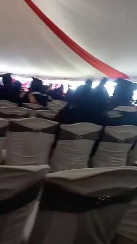 The video shows a graduation ceremony taking place under a white tent. The tent is decorated with a red banner. Rows of chairs covered in white fabric and accented with gray ribbons and buckles are visible. A group of people wearing graduation gowns are standing or sitting. Some are interacting and gesturing. The floor appears to be grass.