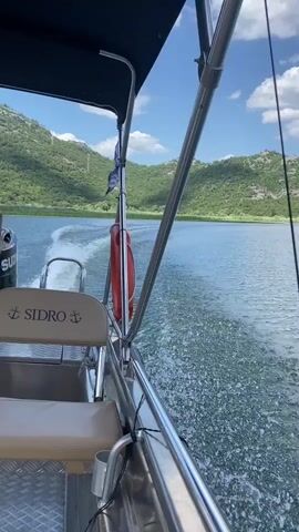 The video shows a view from a moving boat on a body of water. The boat has a metal railing and a black canopy. A red life preserver is visible hanging on a pole. The water is choppy, with a white wake trailing behind the boat. In the distance, lush green, tree-covered hills rise under a blue sky with scattered clouds. The words "SIDRO" and an anchor symbol are visible on the boat's seat. The perspective is from the right side of the boat, looking forward and slightly to the right.