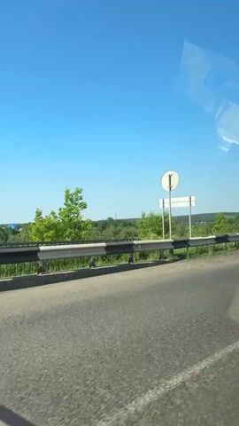 The video shows a view from a moving vehicle on a paved road. A metal guardrail with alternating black and white sections runs along the right side of the road. Beyond the guardrail, there is a grassy area and then a landscape with trees and some buildings visible in the distance. The sky is clear and blue. In the middle of the video, a silver SUV passes the vehicle from which the video is being recorded. The SUV has tinted windows and a roof rack. A traffic light pole is visible on the right side of the road. The video appears to have been taken on a sunny day.