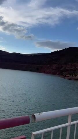 The video shows a body of water from the perspective of someone standing on a bridge. The water is green-blue and has small waves. In the distance, there are mountains and hills with reddish-brown soil. The sky is cloudy with patches of blue sky and sunlight. The bridge has a concrete surface and a railing with pink and white bars. There is a strong wind blowing.