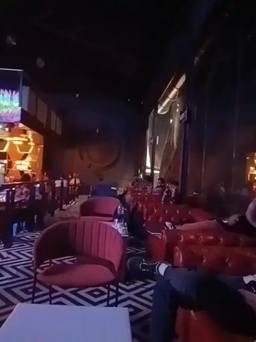 The video shows the interior of a restaurant or bar, with people seated at tables, talking, and walking around. There is a bar area with stools, as well as a separate dining area. The interior is dimly lit, with decorative lighting fixtures, including a large chandelier. There are also TV screens showing various images. The overall atmosphere seems lively. One person says "10".