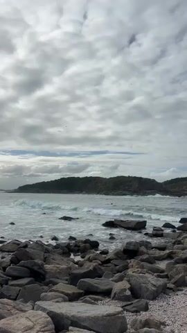 The video shows a daytime scene of a rocky coastline. In the foreground, numerous grey and brown rocks of various sizes are scattered along the shore and partially submerged in the water. Waves are actively breaking against these rocks and rolling towards the shore, creating white foam. Further out in the water, two individuals can be seen, appearing to be surfers or swimmers, navigating the waves.

In the mid-ground, the ocean stretches out, with visible waves. The background features a prominent, green-covered rocky headland or small island. The sky is overcast with a mix of light and dark grey clouds, but the overall lighting suggests it is daytime. No specific time of day beyond daytime can be inferred. The weather appears cloudy but not stormy. The natural environment suggests a coastal area, specifically Cabo Frio, Brazil, based on the provided context. There is no visible text in the frame. The audio contains Spanish speech, indicating a conversation is taking place off-camera or about elements within the scene.