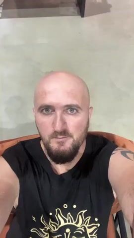 A bald man with a beard and green eyes is sitting in an orange chair, wearing a black t-shirt with a sun design. He is looking at the camera. The background appears to be an indoor seating area with orange couches and a concrete floor. There is a table behind him. The lighting is dim.