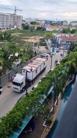 A white semi-truck with a long white cargo trailer drives down a paved road lined with lush green trees and bushes. On top of the trailer, two people in red shirts are standing. A person in a white shirt is standing behind the truck. Several other people and motorcycles are visible on the road and sidewalk further down. In the background, there are multi-story buildings, some under construction, and other residential structures. A swimming pool is visible on the right side of the frame, with blue water. The sky is overcast.