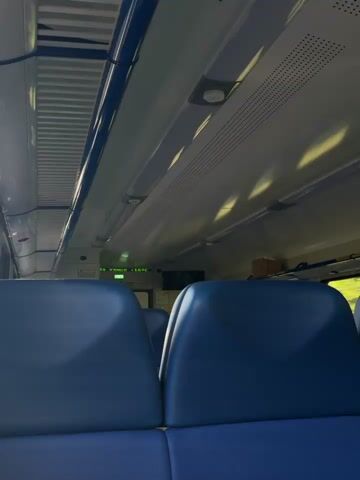 The video pans across the interior of a train carriage. Two blue seats are visible in the foreground, facing away from the camera. Above the seats, fluorescent lights illuminate the ceiling of the carriage. A digital display shows "16°C" which indicates the temperature inside the carriage. To the right of the display, a small television screen is mounted on the wall. In the distance, luggage is stored on an overhead rack. The overall environment appears to be clean and well-maintained.