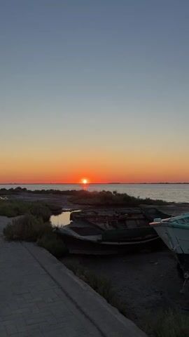 This is a vertical, short video taken during sunset. The sun is a small orange orb, low on the horizon, casting a warm glow on the water and sky. The sky transitions from a pale blue at the top to a fiery orange near the horizon. In the foreground, there are several old, weathered boats resting on the muddy shore. To the left, there's a paved walkway and some sparse vegetation. The overall scene is tranquil and evokes a sense of calm and natural beauty.