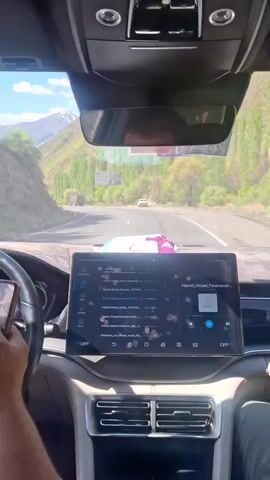 Here is a description of the video.
A person is driving a car, enjoying the music of Hamid Hirad, and looking out at the scenery. The road is winding through the mountains, and there are trees on either side. A military-style truck with a green tarp is parked on the side of the road. The interior of the car is modern, with a large tablet in the center console.  The weather is sunny.