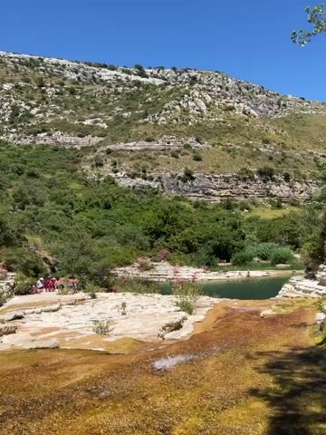 The video shows a natural landscape with a body of water, possibly a river or a small lake, in the foreground. The water has a warm, orange hue in some areas and a deeper green in others. There are rocky, uneven surfaces around the water. In the mid-ground, there is a lush green area with trees and some pink flowers. A group of people, appearing to be mostly women and children, are sitting and standing in this area. Some of them are wearing bright clothing. Further back, there are more trees and vegetation. In the background, a large, rocky hill or mountain dominates the scene, with layers of rock visible. The sky is clear and blue. The overall impression is a sunny day in a natural, possibly rural or mountainous, environment.